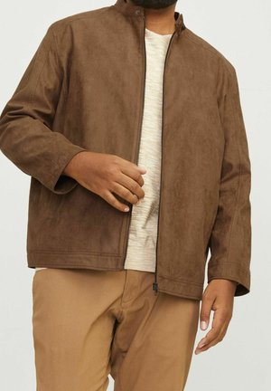Homme portant une veste zippée marron sur une chemise beige clair et un pantalon couleur sable sur un fond uni.