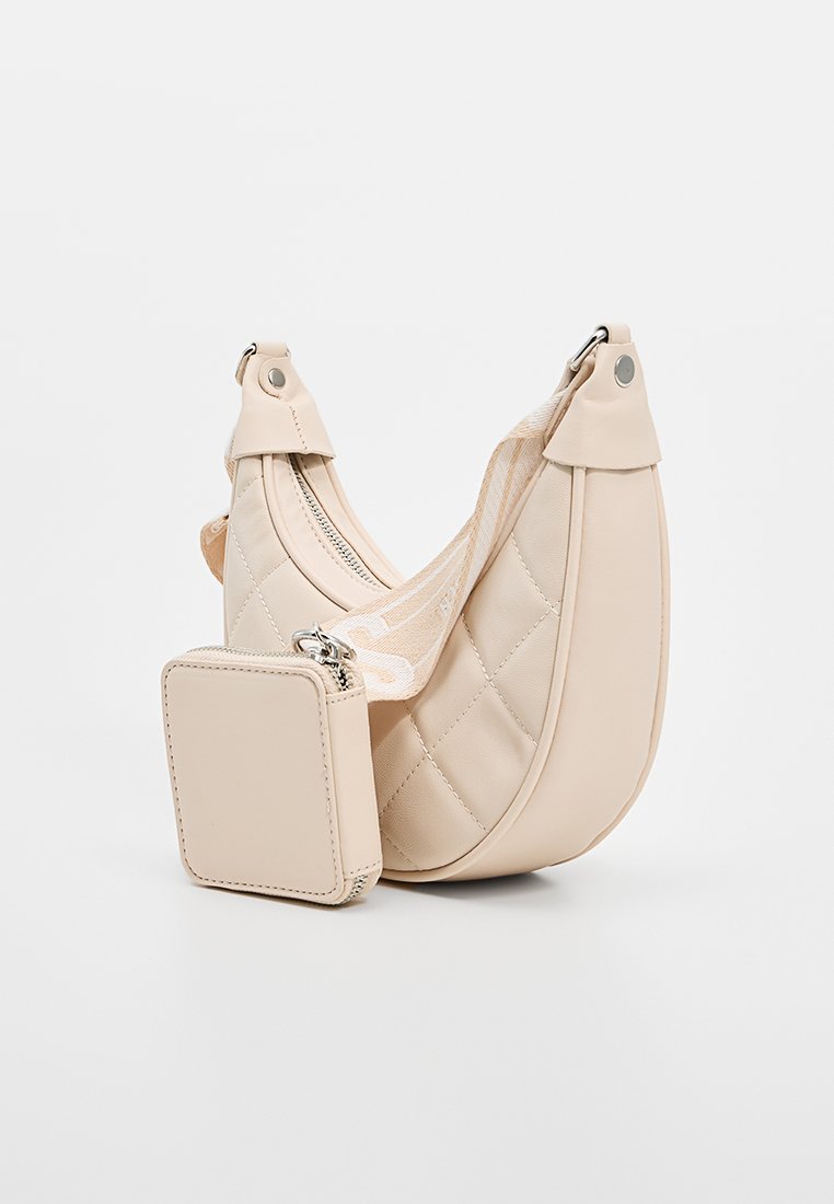Beige Schultertasche aus Kunstleder mit einem gesteppten Design, verstellbarem Tragegurt und einem kleinen abnehmbaren Reißverschlussbeutel, der angebracht ist.