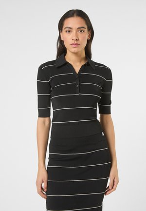 Kobieta w czarnej, krótkim rękawem, koszulowej bluzce z kołnierzykiem oraz dopasowanej spódnicy z cienkimi, białymi, poziomymi paskami, stojąca na jednolitym tle.