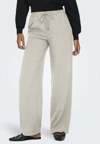 Pantaloni beige a gamba larga con vita elasticizzata e cordoncino, indossati con ballerine nere a punta con borchie e un top nero a maniche lunghe.