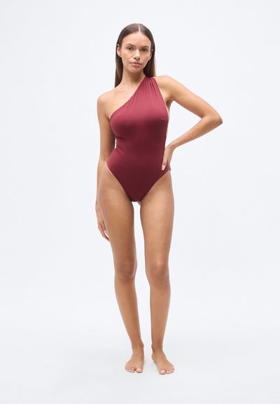 Μπορντό ολόσωμο μαγιό με ένα λουράκι στον έναν ώμο, λεία υφή, κοψίματα στα μπούτια και μινιμαλιστικό σχεδιασμό.