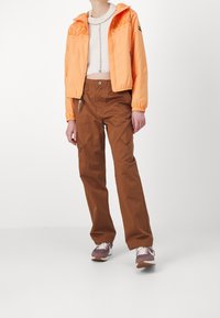 Pomarańczowa lekką kurtka z elastycznymi mankietami, w połączeniu z brązowymi spodniami cargo z bocznymi kieszeniami, noszona z fioletowo-białymi trampkami.