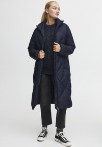 Lang marineblå polstret frakk med høy krage og quiltet design, brukt over en mørk strikket genser og lysegrå avkuttede jeans, kombinert med svarte joggesko.