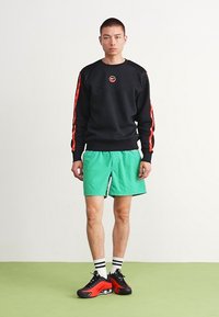 Sort sweatshirt med røde detaljer, grønne shorts og sorte/røde sneakers. Modellen står på et lyst grønt gulv, med armene afslappet langs siderne.