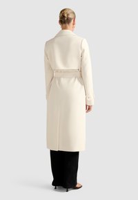 Frau mit einem blonden Haarknoten, die einen langen cremfarbenen Trenchcoat, schwarze Hose und offene High Heels trägt, steht mit dem Rücken zu einem schlichten Hintergrund.