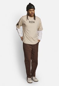 Beige t-shirt met zwarte tekst en logo-accenten, gelaagd over een witte lange mouw shirt. Bruine broek en witte sneakers maken de look compleet.