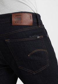 Ciemnoniebieskie dżinsy z denimu z skórzaną naszywką marki oznaczoną jako "RAW" oraz pomarańczowym przeszyciem. Posiadają standardowy design tylnych kieszeni.