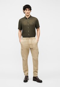 Άνδρας που φοράει σκούρο πράσινο πουκάμισο με κοντά μανίκια και κουμπιά, μπεζ παντελόνι cargo και σκούρα καφέ μπότες, στέκεται με τα χέρια στις τσέπες.