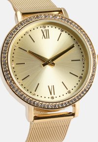 Orologio color oro con cinturino in rete testurizzata. Cassa rotonda con quadrante dorato metallico, numeri romani e una lunetta incastonata di cristalli.