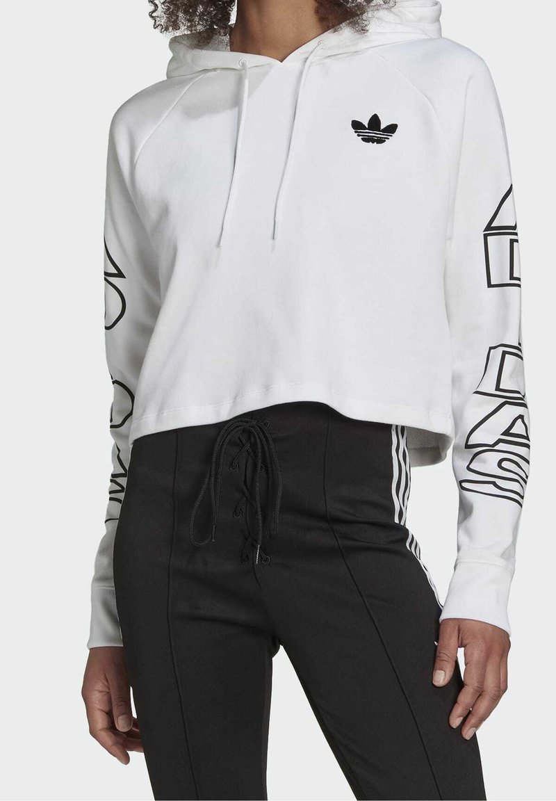 White cropped hoodie with black Adidas logo; features drawstrings and bold black text down the sleeves. Paired with black fitted pants.