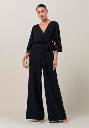 Žena v čiernom zavinovacom overale so širokými nohavicami, držiaca béžové puzdro, obutá v zlatých sandáloch a so zavesenými náušnicami.