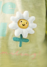 Flor de tela suave con pétalos blancos, una cara amarilla sonriente y tallo verde cosido sobre un fondo verde claro con texto parcial.