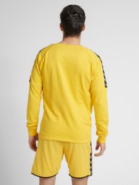 Gul langærmet skjorte med rund halsudskæring og sorte accentstriber på skuldrene. Matchet med gule shorts i samme farve med sorte kanter.