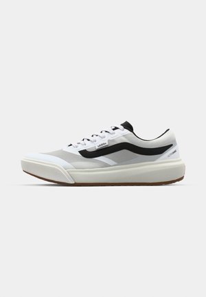 Sneaker bianca e grigia con accenti neri, caratterizzata da una suola spessa e un design con lacci, esposta su uno sfondo chiaro e semplice.