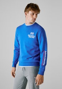 Νέος άντρας φορώντας μια μπλε sweatshirt της Red Bull Racing και γκρι φόρμες, κοιτάζοντας αριστερά σε έναν απλό φόντο.