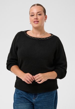 Femme portant un pull noir à manches longues avec un col clouté et un jean bleu, debout, les mains doucement jointes devant elle.