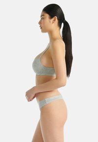 Frau mit langen dunklen Haaren in einem Pferdeschwanz, die einen hellgrauen BH und passende Unterwäsche trägt, im Profil vor einem einfarbigen Hintergrund stehend.