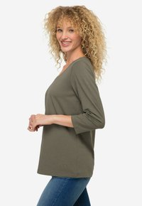 Oliwkowa bluzka z długim rękawem, dekoltem w serek, luźny krój oraz haftowany logo na dole. Miękka tkanina i prosty, elegancki design.