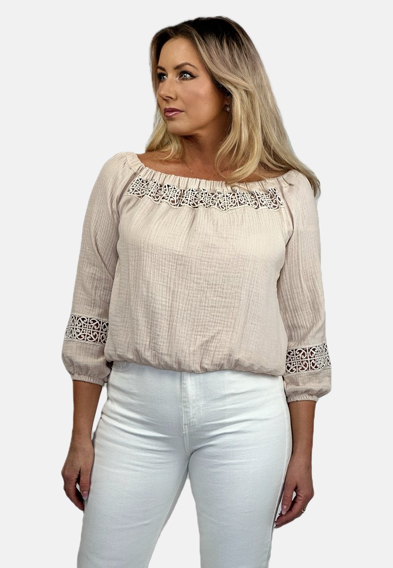 Jasnoróżowa bawełniana bluzka o teksturowanym wykończeniu, z dekoltem odsłaniającym ramiona oraz koronkowymi zdobieniami na rękawach i u dołu. Połączona z białymi spodniami.