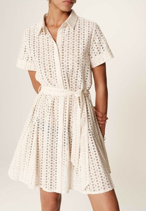 Robe en dentelle blanche avec un col, des manches courtes et une taille cintrée. Le tissu présente des motifs découpés circulaires et une jupe évasée.