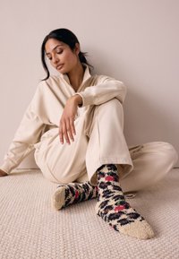 Chaussettes douillettes aux motifs marron et crème, présentant une texture duveteuse avec des touches de rouge et de vert, portées avec une tenue décontractée beige sur un tapis texturé.