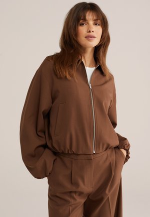 Donna che indossa una giacca marrone con cerniera e orlo elasticizzato e pantaloni marroni coordinati, in piedi con le mani nelle tasche su uno sfondo beige.