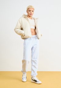 Béžová péřová bunda s kapucí, krémový crop top, světle modré tepláky a bílé tenisky s černými akcenty. Hladké, jemné textury.