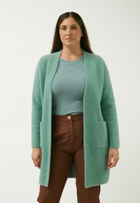 Aquamarin-fuzzy cardigan med åben front, to forlommer og lange ærmer, lagd over en struktureret lyseblå sweater og brune bukser.