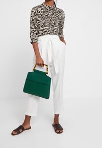 Zelená strukturovaná kabelka s rukojetí z želvoviny, ve spojení s béžovou košilí se zvířecím potiskem a bílými kalhotami s vysokým pasem, nošená s hnědými sandály.