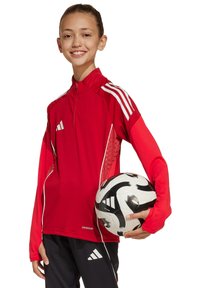 Rode sportieve longsleeve top met een halve rits en witte strepen, gecombineerd met zwarte shorts en houdt een zwart-wit voetbal vast.