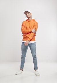 Sudadera con capucha naranja, jeans ajustados de mezclilla azul claro, zapatillas blancas y una gorra blanca. Tejido suave, diseño casual, estilo ajustado.