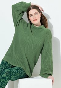Maglione verde lavorato a maglia con collo alto e maniche texture, abbinato a leggings verdi con motivo a foglie, su uno sfondo bianco.
