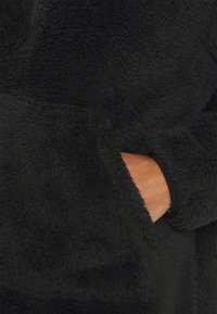 Pull noir en polaire avec un extérieur doux et texturé, et une poche latérale avec une main visible. Fermeture éclair au niveau du cou.