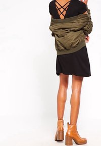 Zielona oliwkowa bomberka noszona na czarnej sukience z krzyżującym się tyłem. Beżowe botki na obcasie sięgające kostki. Gładkie materiały i swobodny styl.