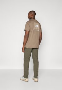 Brun bomull t-skjorte med logo på baksiden, parret med olivengrønne bukser og lyse sneakers. Enkelt, uformelt design.