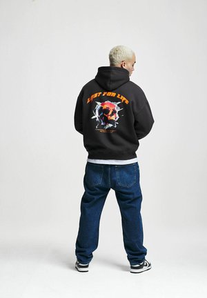 Hoodie in dark gray with front kangaroo pocket and back graphic of a skull surrounded by text. Paired with blue jeans and black sneakers.