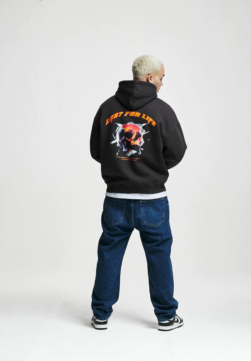 Hoodie in dark gray with front kangaroo pocket and back graphic of a skull surrounded by text. Paired with blue jeans and black sneakers.