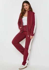 Veste zippée bordeaux et pantalon de survêtement assorti avec des rayures blanches sur les côtés. Tissu lisse, coupe décontractée, associé à des baskets de couleur claire.