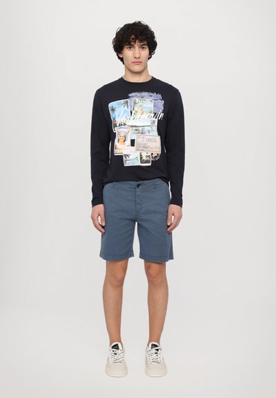 Camisa de manga larga azul marino con diseño de postales gráficas; combinada con pantalones cortos azul claro; modelo lleva zapatillas blancas.