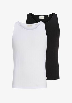 Jeune homme portant des lunettes de soleil rondes, un débardeur blanc, une ceinture noire, un jean noir ample et des chaussures noires, debout devant un fond clair uni.