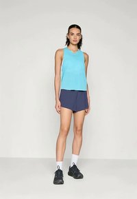 Γυναίκα που στέκεται φορώντας τοπ χωρίς μανίκια σε τουρκουάζ χρώμα, ναυτικές σορτς, λευκές κάλτσες και μαύρα αθλητικά παπούτσια μπροστά σε απλό φωτεινό φόντο.