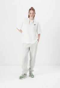 Balta per didelė t-shirt su žaliu logotipu, derinama su pilkais sportiniais džinsais. Modelis dėvi žalias ir pilkas sportines batus, stovi prieš baltą foną.