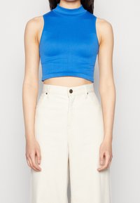 Kvinna som bär en ärmlös blå crop top och högmidjade vita vida byxor mot en enkel bakgrund.