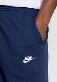 Mano en el bolsillo de unos pantalones deportivos Nike azul marino con logo blanco, parte de una camiseta blanca visible por encima de la cintura.