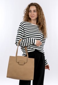 Sac fourre-tout en cuir beige avec une forme structurée, une poignée supérieure et une fermeture à rabat. Présente des coutures visibles et une surface texturée.