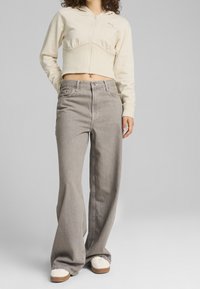Beige cropped hoodie met ritssluiting en capuchon, voorzien van een tonale logo, gecombineerd met lichtgrijze wijde jeans en witte sneakers met bruine accenten.
