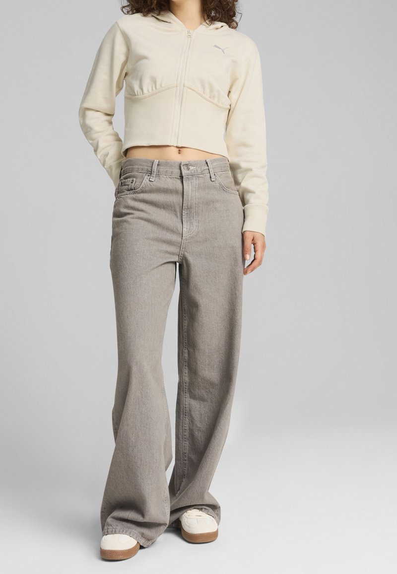 Beige cropped hoodie met ritssluiting en capuchon, voorzien van een tonale logo, gecombineerd met lichtgrijze wijde jeans en witte sneakers met bruine accenten.