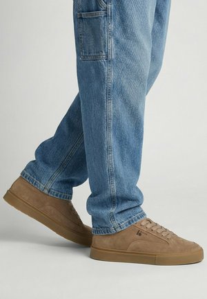Person, die hellblaue Jeans und beige Wildledersneaker trägt, steht mit einem leicht angehobenen Fuß.