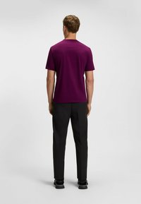 Lila kortärmad t-shirt med rund hals, parat med svarta byxor och svarta skor, sedd bakifrån mot en neutral bakgrund.