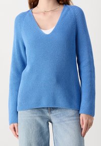 Woman wearing a bright blue ribbed V-neck sweater over a white undershirt and light blue jeans, standing against a plain background.